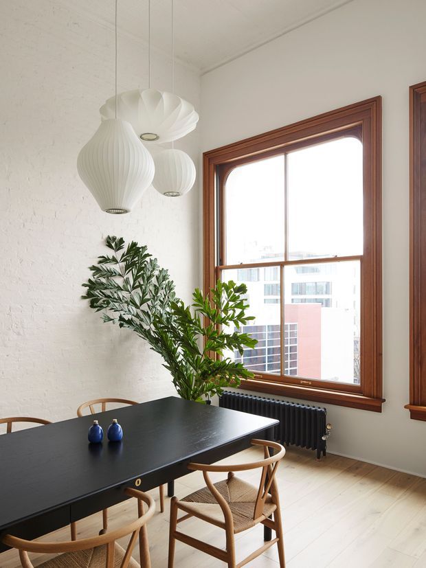 Bright dining area with a large black rectangular dining table, timber-framed windows, light timber flooring, exposed white-painted brick walls, a black radiator, and sculptural white pendant lights. Wooden wishbone-style chairs and a tall leafy plant add warmth and contrast. The space has a refined modern Scandinavian character with traditional timber window detailing.