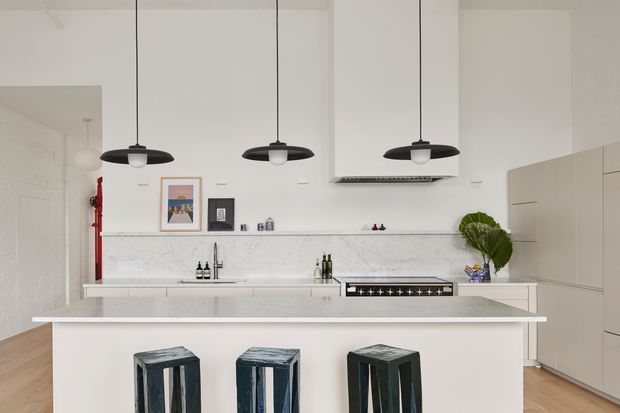 Bright contemporary kitchen with a large white island, three dark metal pendant lights, pale marble-look splashback and benchtops, integrated cabinetry, a freestanding-style cooktop, and three dark stools. Light timber flooring and minimal decorative elements create a clean, refined interior.
