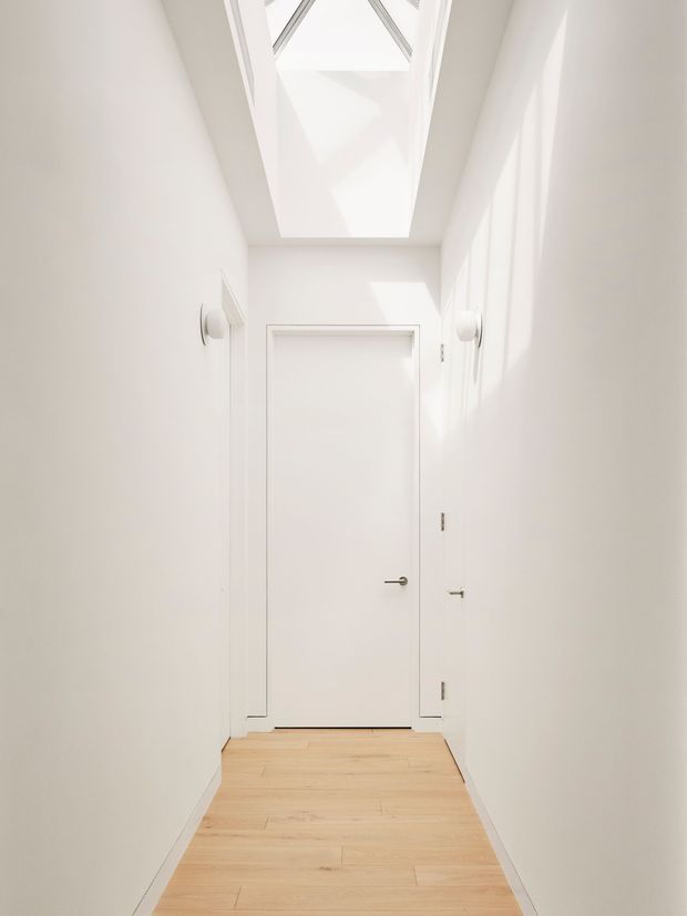 A narrow, minimalist white hallway with a flush white door at the end, pale timber flooring, concealed or minimal trim, and a large overhead skylight bringing in bright natural light. Simple white wall-mounted fixtures and understated hardware reinforce the clean architectural character.