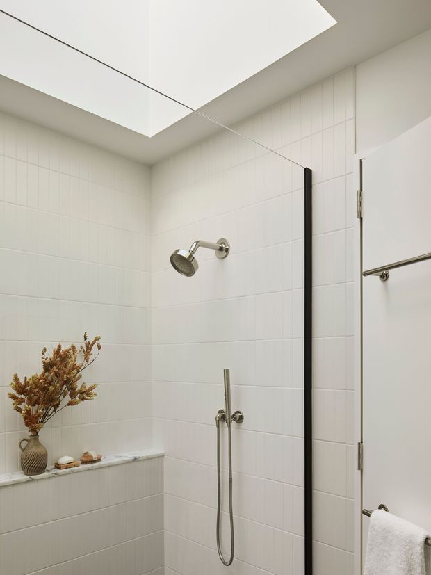 Bright, minimalist bathroom shower with white vertically lined wall tiles, a large frameless skylight or ceiling opening, brushed metal shower fixtures, a slim black shower screen frame, and a white towel hanging on the door. A small built-in tiled ledge holds a vase of dried botanicals and toiletries.