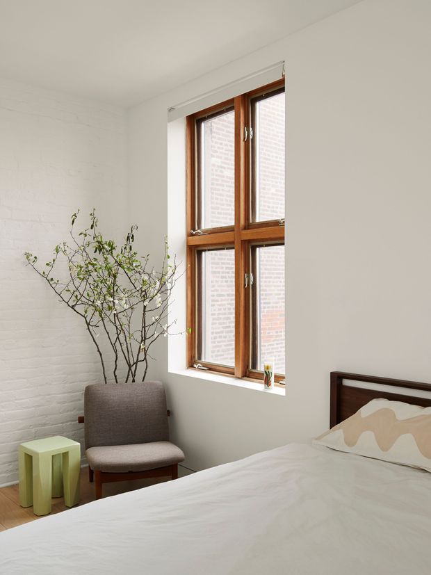 Bright, minimalist bedroom with white-painted brick walls, a warm timber-framed divided window, and a simple bed with a dark timber headboard and neutral bedding. A compact upholstered chair, small pale green side table, and a sparse branch arrangement add understated natural accents.