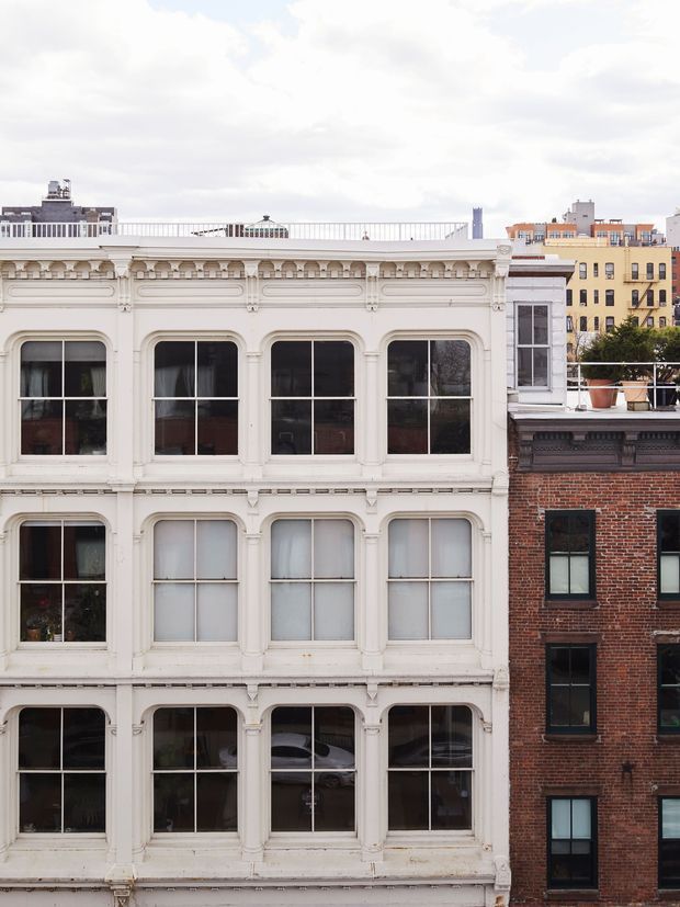 Street-facing view of a multi-storey historic urban building with an ornate white-painted facade, tall vertically proportioned windows, decorative mouldings, cornices and pilasters. A contrasting exposed red-brick building stands adjacent on the right, with additional city buildings and rooftop terraces visible in the background under an overcast sky.