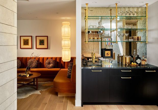 A sophisticated open-plan entertaining area featuring a dark built-in bar with brass handles, mirrored backsplash, and glass shelving supported by brass fittings. The adjacent lounge has a rich rust-orange upholstered banquette and curved seating, patterned cushions, framed artwork, a sculptural pendant light, and timber flooring. The overall composition is warm, elegant, and distinctly Art Deco-inspired.