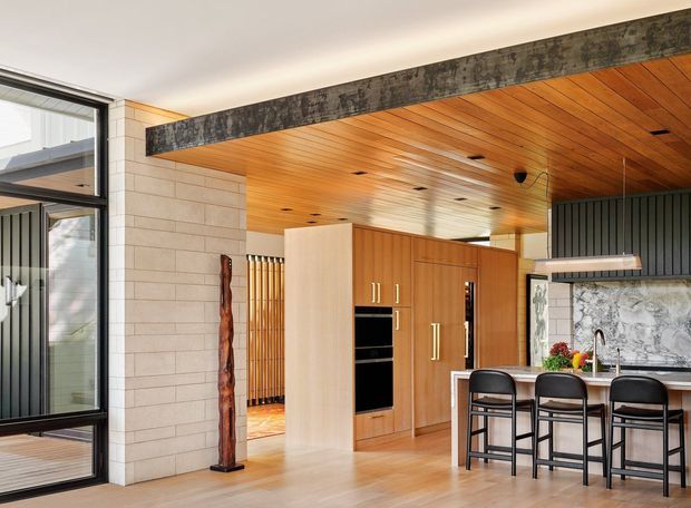 Bright contemporary kitchen and dining area with extensive warm timber ceiling and fixed timber cabinetry, a large stone island with bar seating, and a pale masonry feature wall. Full-height glazing and black-framed windows open to an exterior courtyard or deck. A dark exposed structural beam spans the ceiling, while a sculptural timber screen and vertical slats define the adjacent circulation area.