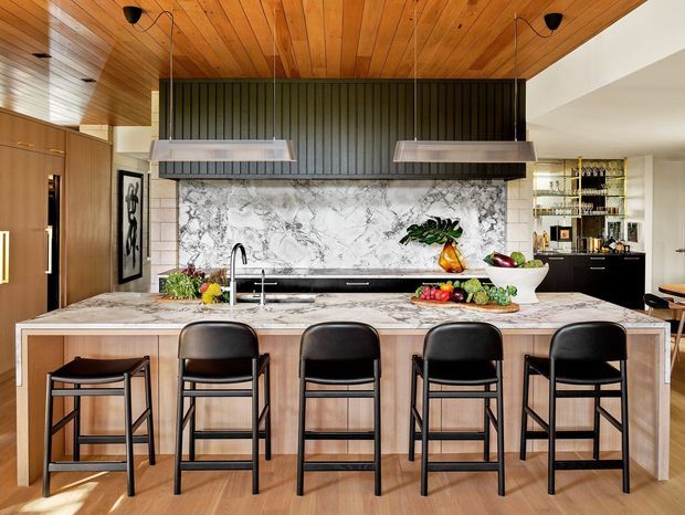 A spacious contemporary kitchen centred on a large island with a pale marble-look surface, integrated sink and four black bar stools. Warm timber cabinetry and a timber-lined ceiling contrast with a dramatic veined stone splashback, dark vertical feature panelling, twin stainless-steel rangehoods and black-framed fixtures. A compact drinks or bar area with open shelving is visible in the background.
