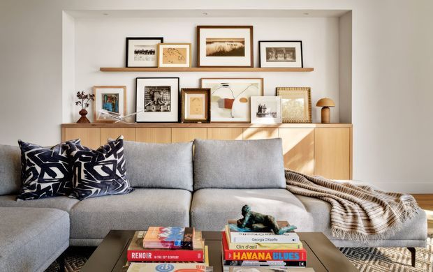 A warmly lit contemporary living room featuring a large light-grey sectional sofa with patterned navy-and-white cushions and a draped textured throw. A low coffee table holds stacked books and a small sculpture. Behind the sofa is a long timber credenza with framed artwork and photographs arranged on top and on a floating timber picture ledge, set within a recessed wall niche.