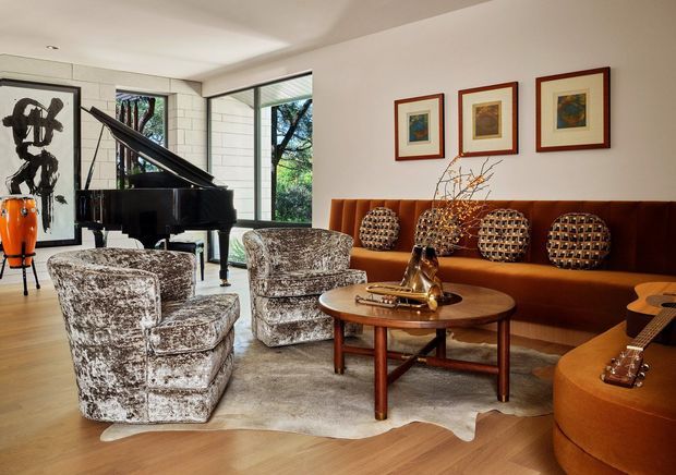 A richly furnished living room with a grand piano, sculptural patterned lounge chairs, a long rust-coloured upholstered sofa, round wooden coffee table, framed artwork, and a guitar in the foreground. Large windows bring in natural light and overlook greenery. The space has warm timber flooring, a neutral ceiling and walls, and layered vintage-inspired furnishings.