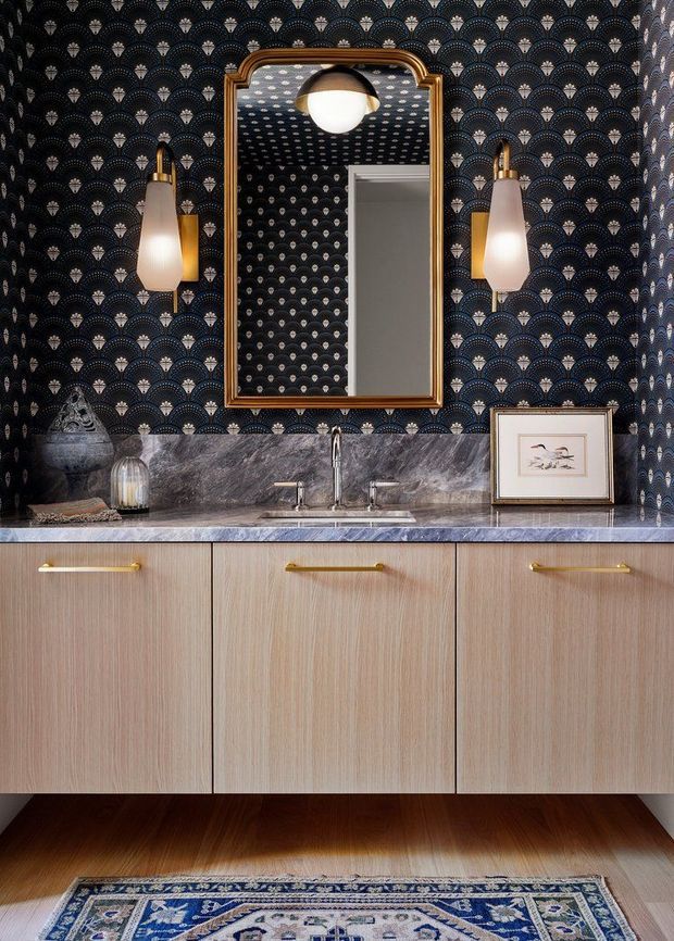 A richly detailed powder room vanity with pale timber floating cabinetry, a dark veined stone benchtop and integrated basin, framed mirror, and twin wall sconces. Dramatic fan-pattern wallpaper covers the walls and ceiling, complemented by brass fixtures, a framed artwork, dark timber flooring, and a patterned rug. The styling is bold and decorative with strong Art Deco influence.