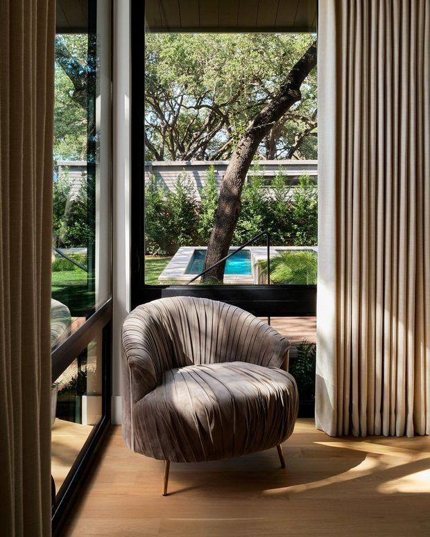 A sunlit interior corner with a sculptural upholstered lounge chair positioned beside full-height black-framed glazing. Floor-to-ceiling curtains frame views of a landscaped garden and swimming pool, while warm timber flooring and strong natural light create a refined, intimate sitting space.