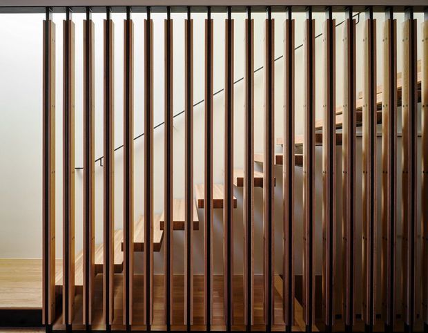 Interior view of a contemporary stairway screened by closely spaced vertical timber slats. A timber staircase rises through the centre, with warm wood flooring and pale walls; dark metal or painted framing creates a strong linear rhythm and partially conceals the upper level.