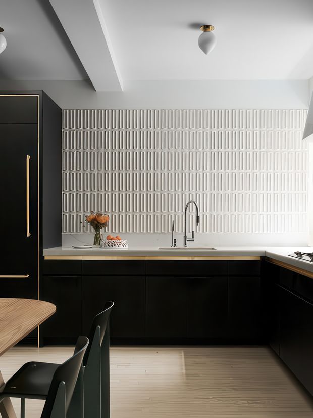 Contemporary kitchen with a dramatic black-and-white palette. Matte black full-height cabinetry and lower joinery are accented with slim brass-gold handles and trim, while a white benchtop and sculptural white textured tile backsplash create a strong focal wall. A dining table and dark chairs occupy the foreground, and a streamlined sink with a tall gooseneck tap sits beneath the backsplash. Recessed ceiling detailing and small pendant fixtures add refined architectural character.