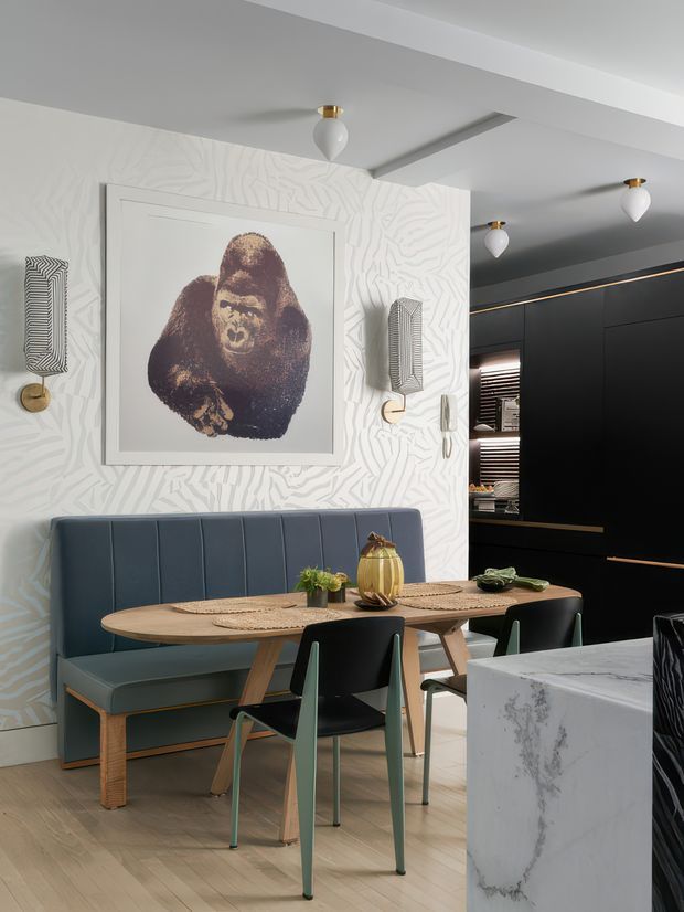 A refined contemporary dining area with a built-in blue upholstered banquette, sculptural timber dining table, and contrasting dark dining chairs. The space features a white textured patterned wall with framed gorilla artwork, decorative wall lights, pale timber flooring, and a dramatic black built-in kitchen or storage wall with brass detailing. A white marble-look island or counter occupies the foreground.
