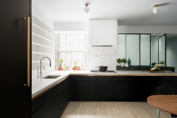 A bright contemporary kitchen with a U-shaped layout, matte black lower cabinetry, white stone benchtops, a white marble-look splashback, and a large central rangehood. The room features a black-framed internal glass partition or window, a white grid-pattern feature wall, timber-toned flooring, brass hardware, indoor plants, and a round timber dining table.