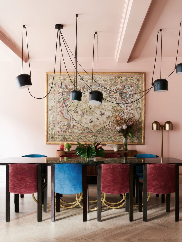 A richly styled dining room with a long dark dining table surrounded by upholstered chairs in burgundy, vivid blue, and teal. The room has pale blush-pink walls and ceiling with decorative trim, herringbone timber flooring, a large ornate framed tapestry or artwork, sculptural black pendant lights with exposed cords, brass floor lighting, and colourful floral arrangements. The overall design is eclectic, bold, and refined.