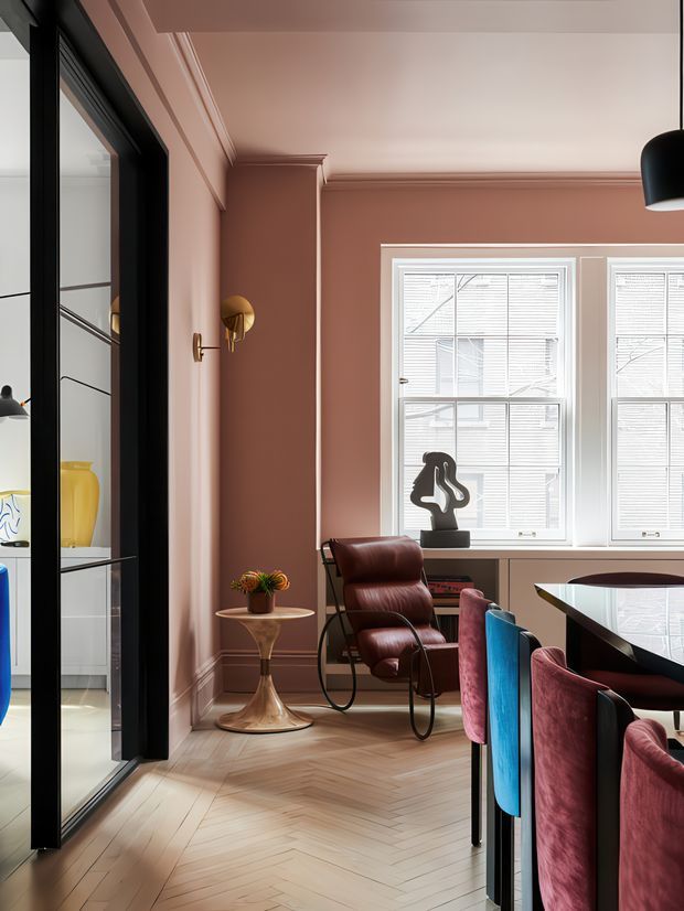 A refined dining area with dusty rose walls, white-trimmed windows and blinds, and a pale timber herringbone floor. A dark contemporary dining table with jewel-toned upholstered chairs occupies the foreground, while a sculptural leather lounge chair, brass wall sconce, small side table, and pendant light add layered detail. Black-framed glazing opens toward a bright adjacent room with built-in cabinetry. The space blends classic architectural mouldings with bold contemporary furnishings and warm, sophisticated colours.