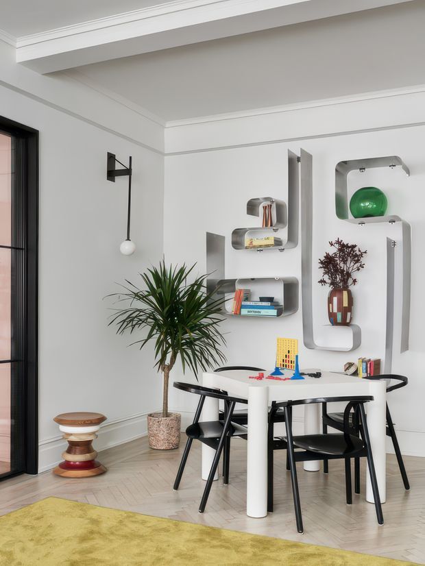A refined dining and workspace corner in a bright room with white decorative wall mouldings, a black-framed glazed door or window, sculptural white wall shelving, and a round white table surrounded by black chairs. Books, small objects, and a green glass vessel decorate the shelving, while a tall indoor plant and colourful sculptural side stool add visual interest. Pale herringbone timber flooring and a yellow-green rug create contrast.