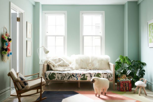 A bright, playful living room with pale green walls, tall white-trimmed windows, and a patterned upholstered sofa layered with cream throws and cushions. A mid-century-style wooden armchair sits at the left, while abundant indoor plants, colourful artwork, a whimsical sheep-shaped stool, and a multicoloured side table add eclectic character. Sunlight creates strong highlights and shadows across the layered rugs and timber floor.