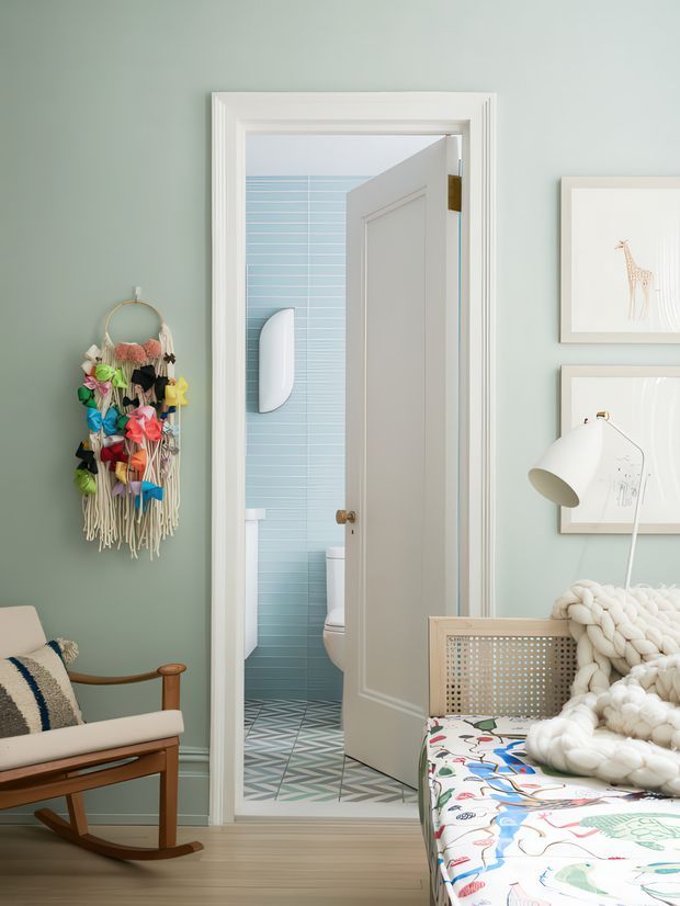 Bright, playful bedroom with a muted sage-green wall, white traditional-style trim, a wooden rocking chair, woven wall hanging, framed artwork, a white floor lamp, and a woven-front bed or daybed covered with a colourful patterned textile and chunky cream knit throw. An open door reveals a compact bathroom with pale blue horizontal wall tiles and patterned geometric floor tiles.