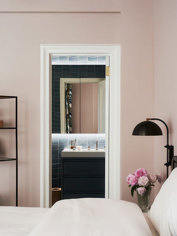 View from a softly styled bedroom toward an open bathroom doorway. The bedroom has blush-pink walls, white bedding, a black arched floor lamp, shelving, and flowers. The bathroom beyond features dark teal tiled walls with pale blue wave-pattern tile bands, a black vanity with a light benchtop, brass-toned fixtures, and a mirrored cabinet. White decorative door trim frames the opening.