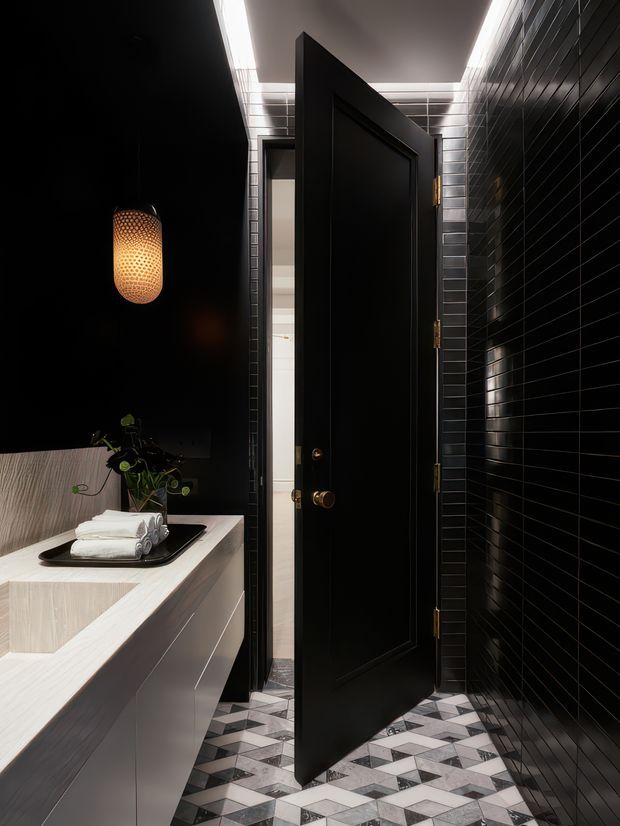 A dramatic, compact bathroom interior viewed toward an open black paneled door. Glossy black rectangular wall tiles cover the right wall and surround the doorway, while a geometric black, white, and grey patterned floor adds contrast. A pale stone or light-toned vanity with integrated basin occupies the left foreground, topped with neatly rolled white towels and a small plant. A warm perforated pendant light glows against the dark wall, and concealed linear ceiling lighting highlights the tiled surfaces.