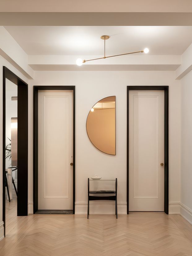 A refined interior hallway with white walls, decorative wall mouldings and ceiling beams, two paneled white doors framed in black, and light timber herringbone flooring. A slim black console table sits beneath a half-round bronze-toned mirror, while a linear brass and globe-bulb ceiling light provides warm illumination. A dark-framed opening at left reveals part of an adjacent room.