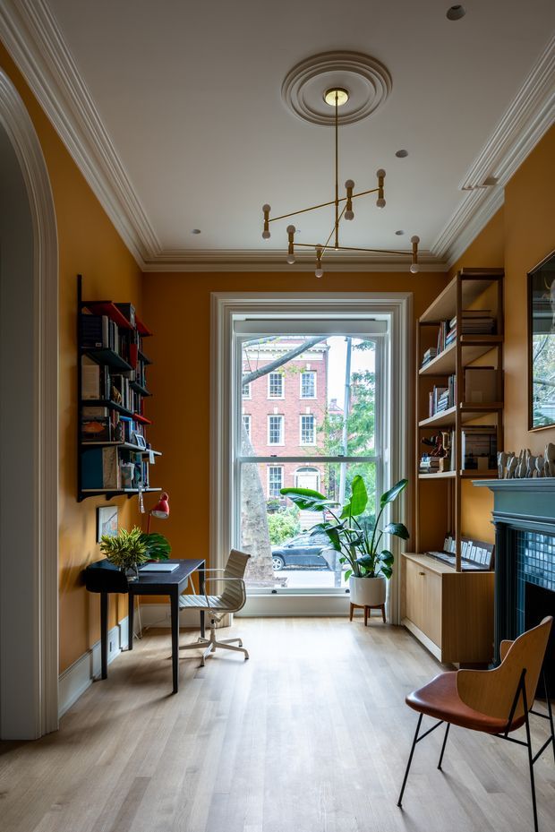 A refined home office in a tall, narrow room with rich ochre-brown walls, pale timber flooring, elaborate white crown moulding and ceiling medallion, and a traditional fireplace surround. Built-in and freestanding open shelving filled with books lines both sides, while a compact desk, ergonomic chair, potted plants, and modern sculptural pendant light create a layered study workspace. A large sash window brings in daylight and views of neighbouring brick buildings and street greenery.