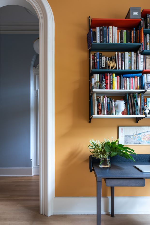 A richly coloured study nook with a dark timber desk, laptop, indoor plant and wall-mounted bookshelves filled with books. The mustard ochre wall is framed by ornate white architectural trim and an arched opening leading to a darker adjoining room with timber flooring. The space combines traditional detailing with bold, curated contemporary furnishings.