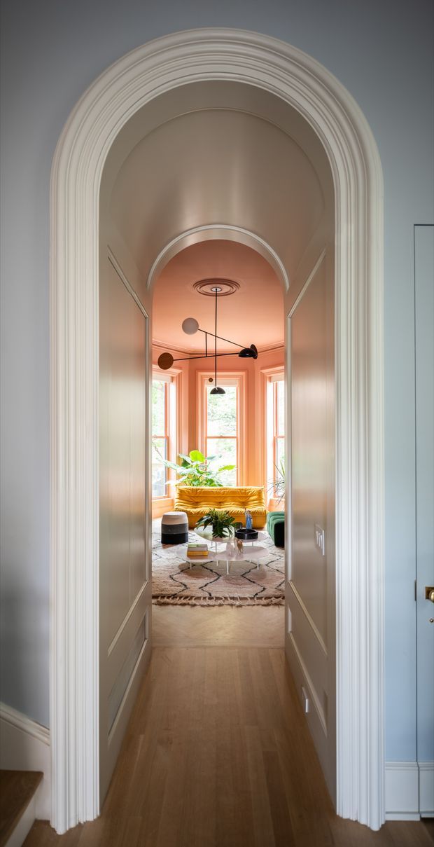 A view through a tall, heavily moulded arched opening and double doors into a bright, richly styled living room. The foreground hallway has pale blue-grey walls, white decorative panelling and trim, and warm timber flooring. Beyond, the living room features a warm coral-pink bay-window surround, a mustard velvet sofa, green seating, abundant indoor plants, a patterned rug, and a sculptural black multi-arm pendant light. The composition highlights the ornate archway, layered cornices, and connection between hallway and living space.