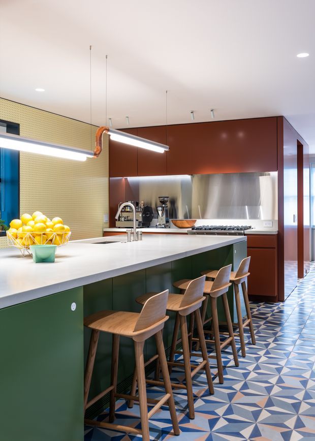 A contemporary kitchen dominated by a large white island with deep green cabinetry and timber bar stools. The kitchen features glossy rust-red overhead cabinetry, a stainless-steel splashback, integrated appliances, a patterned geometric tile floor in blue, white and terracotta, and distinctive linear pendant lighting. A perforated pale yellow wall panel and large windows add texture and light.
