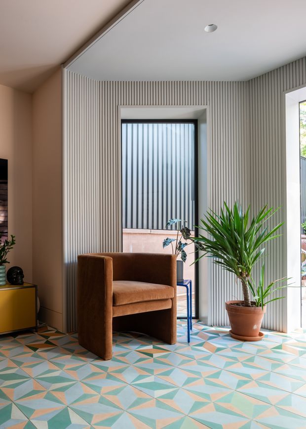 A bright, eclectic sitting area with a sculptural caramel-brown upholstered armchair, a large potted palm, and a low mustard-yellow sideboard. The room features a highly patterned geometric floor in teal, peach, cream, and grey, paired with pale vertical fluted wall panelling and large black-framed windows. Natural light creates an airy yet colourful interior.
