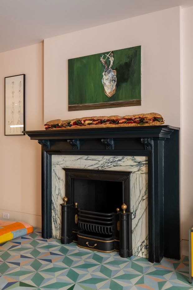 Interior view centered on an ornate black fireplace with classical detailing and a white veined marble surround. A colourful geometric patterned tile floor, pale warm walls, framed artwork, and a sandwich resting on the mantel create an eclectic, character-rich living space.