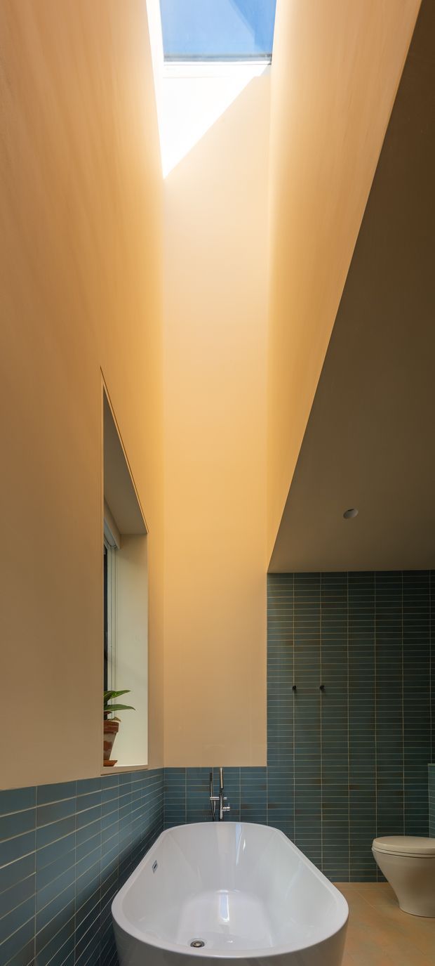 A tall, narrow modern bathroom with a freestanding white bathtub beneath a dramatic skylight. Deep blue-green rectangular tiles line the lower walls and full-height wall behind the bath, while the upper walls are warm pale plaster. A slim vertical window and potted plant appear at left, with a white toilet at right. The space is strongly illuminated by natural sunlight from above.