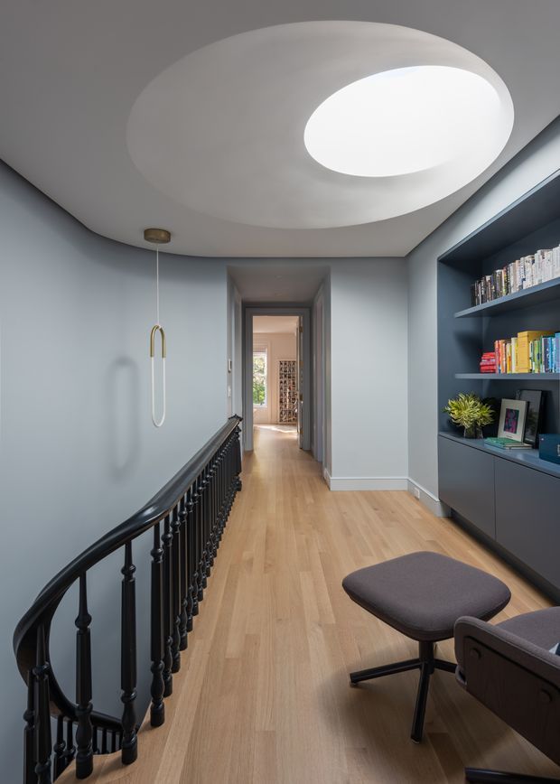 A refined upper-level hallway or landing with pale blue-grey walls, light timber flooring, a sculptural white oval ceiling feature with integrated circular skylight or light well, and a black decorative stair balustrade. A dark built-in shelving and cabinetry unit displays books and small decor on the right. A modern upholstered lounge chair and ottoman sit in the foreground, while a doorway leads to another room in the background.
