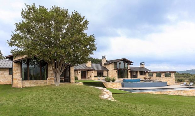 Wide exterior view of a sprawling single-storey residence set in a landscaped lawn, featuring warm tan stone walls, dark metal pitched roofs, large glazed openings, timber detailing, multiple chimneys, and a raised rectangular swimming pool with stone surrounds. Trees and low wooded hills form the background.