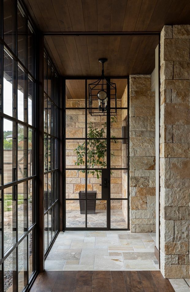 A narrow, refined entry hall or glazed vestibule with dark timber-panelled ceiling, dark timber flooring, black-framed floor-to-ceiling grid windows and a matching glass door. Rough-cut tan and cream stone walls frame the space, while a geometric black metal pendant hangs centrally. A potted tree is visible beyond the glazed door, and natural light enters from the courtyard or exterior.