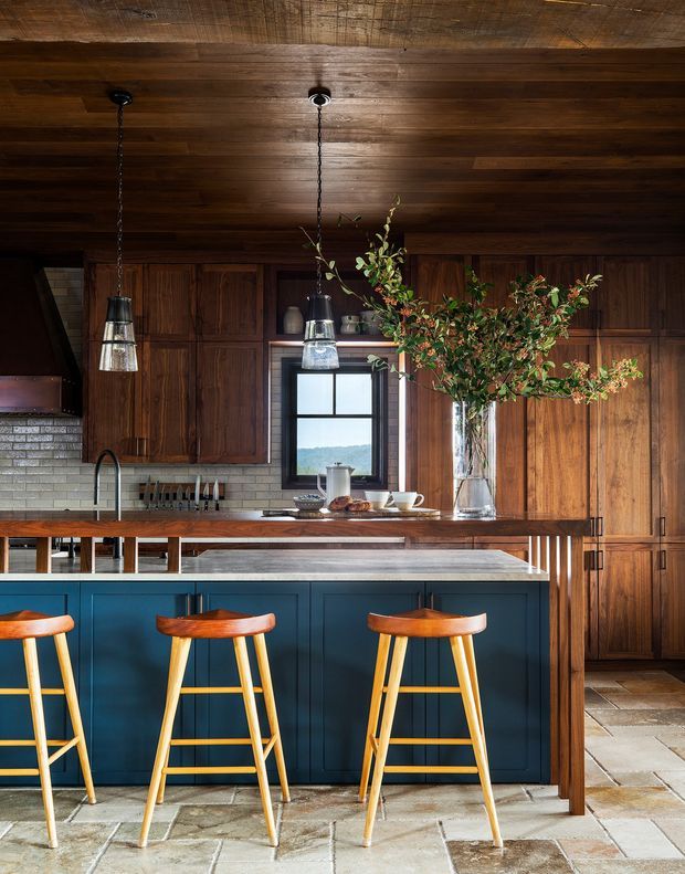 A warmly styled kitchen dominated by dark timber wall and ceiling panelling, with a deep blue island featuring raised timber seating and a wood benchtop. The room includes timber cabinetry, a pale brick-look tiled splashback, pendant lighting, a farmhouse-style sink, and a stone-tile floor. A large vase of branches sits on the island, with a window providing a view of greenery beyond.