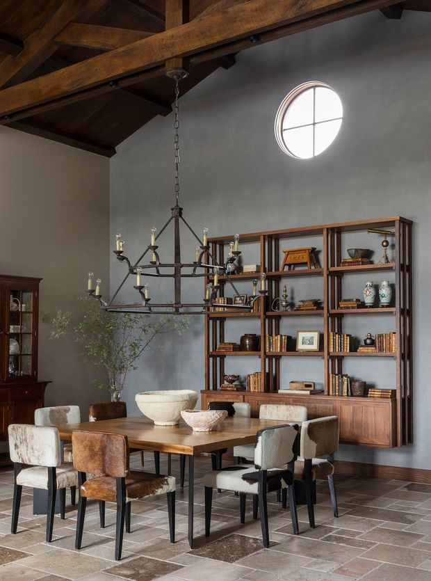 A sophisticated dining room with a large timber dining table surrounded by upholstered chairs, a dark timber built-in bookshelf displaying books and decorative objects, and a vintage-style glass-front cabinet. The room features exposed dark timber roof beams, a grey plastered wall, a circular divided window, a substantial wrought-metal chandelier, and a patterned stone or terracotta tile floor. The overall character is warm, refined, and European-inspired.