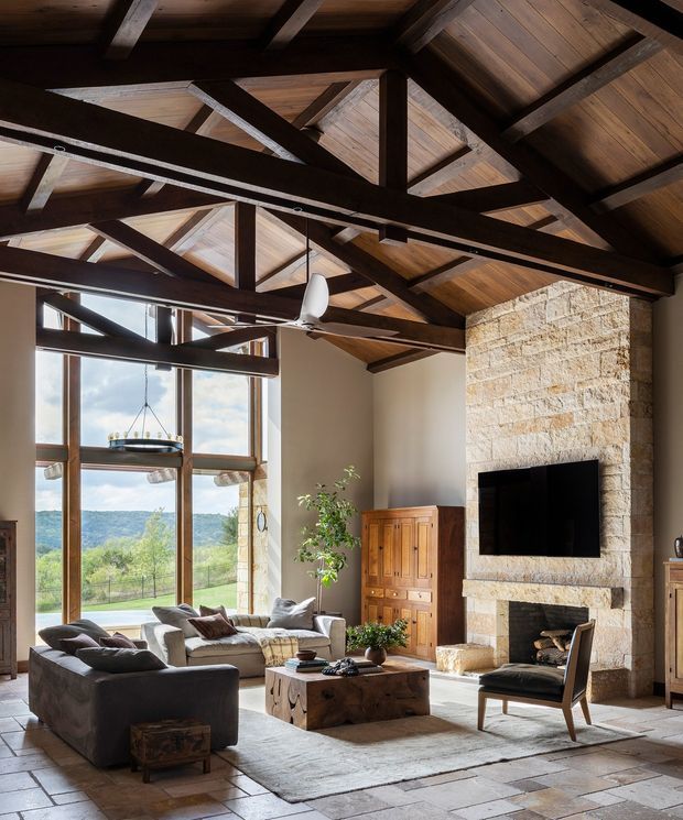 A spacious rustic living room with a dramatic exposed timber post-and-beam ceiling, dark structural trusses, large timber-framed windows, and a tall natural stone fireplace wall with a mounted television. The room includes neutral sofas, a substantial timber coffee table, timber cabinetry, a leather armchair, and a large rug, with a leafy landscape visible through the glazing.