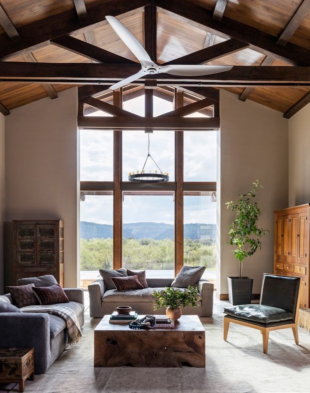A warm, rustic living room with a dramatic timber vaulted ceiling, exposed dark-stained beams and rafters, and a large white ceiling fan. Floor-to-ceiling glazing opens to a scenic landscape and fills the room with natural light. The space includes a sofa, armchair, substantial live-edge timber coffee table, antique-style cabinetry, a potted tree, and a suspended ring pendant light.
