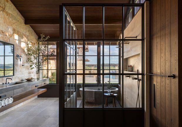 A sophisticated open bathroom suite with a freestanding bathtub enclosed behind a black metal-framed glass partition. The space combines a rough natural stone wall, warm timber ceiling and timber door, large grey stone-look floor tiles, a long floating vanity with dark stone benchtop, and expansive glazing with views to the landscape. The image is photographed from within the bathroom area, with the bathtub and vanity dominating the composition.