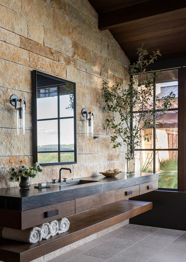 A refined bathroom vanity area with a long floating timber-and-stone console, integrated double basins, dark metal fixtures, wall sconces, and open towel storage. The room features large warm natural-stone wall slabs, dark timber ceiling boards and framing, expansive black-framed glazing, and a neutral stone-tile floor. A landscaped exterior and distant landscape are visible through the windows.