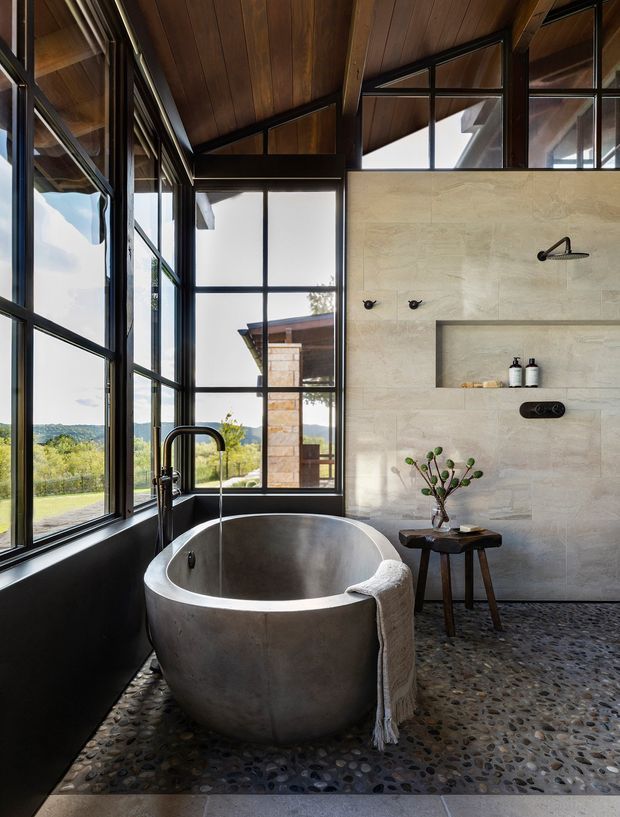 A dramatic, light-filled bathroom with a freestanding concrete bathtub positioned beside full-height black-framed glazing. The room features a pebble mosaic floor, large pale stone wall tiles with a built-in shower niche, exposed dark timber ceiling boards and structural framing, and black plumbing and hardware. A rural landscape is visible through the windows.