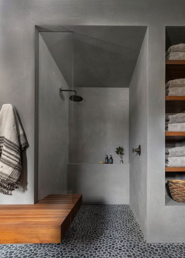 A refined minimalist bathroom with a walk-in shower framed by smooth grey plaster or concrete-look walls. The shower has a black wall-mounted rain shower and recessed ledge, while a pebble mosaic floor extends through the room. Warm timber cabinetry or a built-in bench occupies the foreground, with open timber shelving holding folded towels and baskets on the right. A patterned hanging towel adds texture.