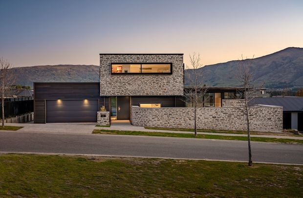 Exterior view of a contemporary two-storey residence at dusk, featuring a prominent rectangular facade clad in coursed grey stone, a long horizontal upper-level window, dark metal cladding, an integrated double garage, and a low stone boundary wall. Mountainous terrain is visible behind the property.