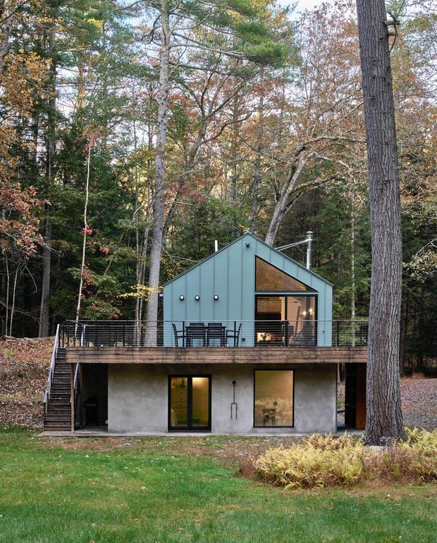 Exterior view of a compact two-storey cabin-like home set within a wooded landscape. The upper level has a steep gabled roof, blue-grey vertical metal cladding, a projecting glazed gable opening, and a timber deck with dark metal balustrade. The lower level has a rough concrete or rendered finish with large glazed doors and windows. Mature deciduous and evergreen trees surround the building.