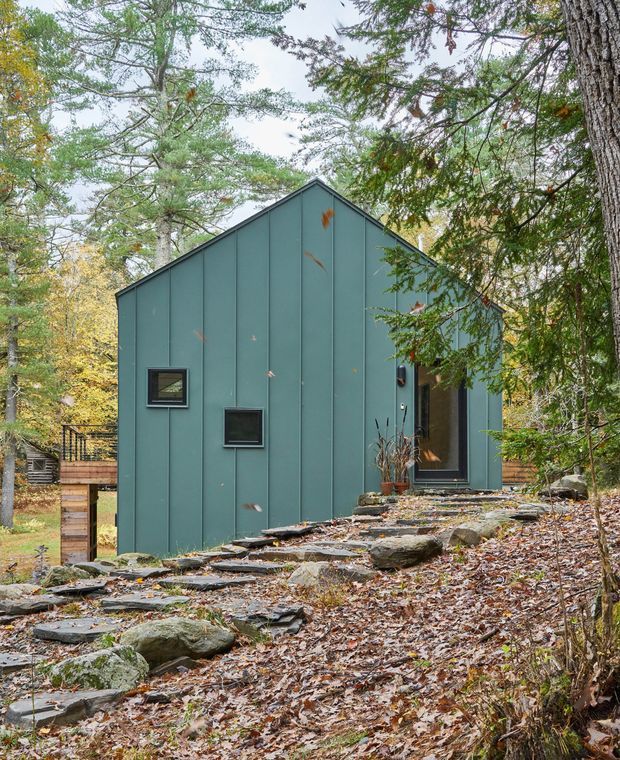 A compact modern cabin-like building set among mature woodland on a sloping, rocky site. The facade has dark teal vertical metal or panel cladding, a simple steep gable roof, small square windows, and a recessed glazed entry. Irregular stone steps lead through leaf-covered ground toward the entrance, with timber fencing and decking partially visible behind the building.