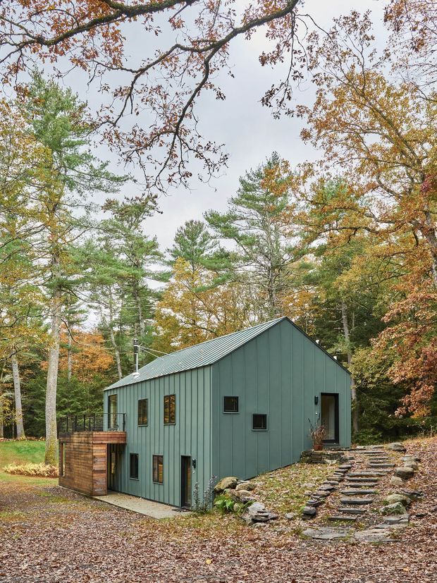 Exterior view of a compact modern cabin set among mature woodland in autumn. The building has a simple gabled form, dark green vertical metal cladding, a standing-seam metal roof, small dark-framed windows, and a timber-clad projecting deck or entry volume. A stepped stone walkway rises through the wooded, sloping site to the entrance.