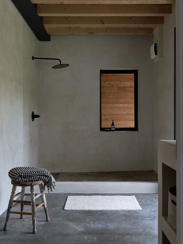 A minimalist walk-in shower bathroom with a raw, plastered finish, concrete floor, exposed timber ceiling beams, and matte black shower fixtures. A small interior window is lined with horizontal timber boards, while a woven stool and bath mat add texture. The space has a restrained, earthy and contemporary aesthetic.