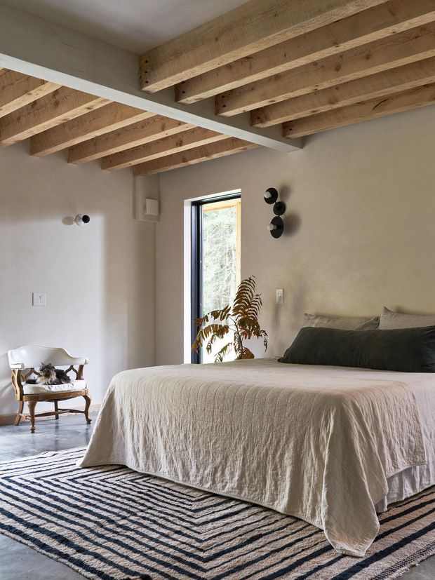 A calm, contemporary bedroom with a low-profile bed dressed in natural linen, dark green-grey pillows, and a striped neutral rug. The room features exposed timber ceiling joists, warm off-white plaster walls, polished concrete flooring, slim dark window framing, black wall sconces, and a vintage-style white armchair. A narrow opening provides views to greenery outside.