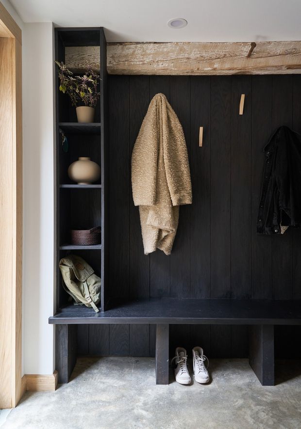 A compact, built-in entryway mudroom with a dark vertical timber-panelled coat and storage unit. The joinery includes open shelving, a long bench, lower shoe storage, and wall hooks holding coats. A large rustic exposed timber beam runs across the upper portion, contrasting with the black joinery. The space has a polished concrete floor, white ceiling and walls, and warm timber trim at the doorway. The styling is practical, earthy, and contemporary with a restrained palette of black, beige, natural wood, and grey.